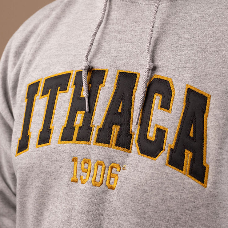 A close-up of a man wearing a heather gray hoodie with 'ITHACA 1906' embroidered in black and gold on the front.