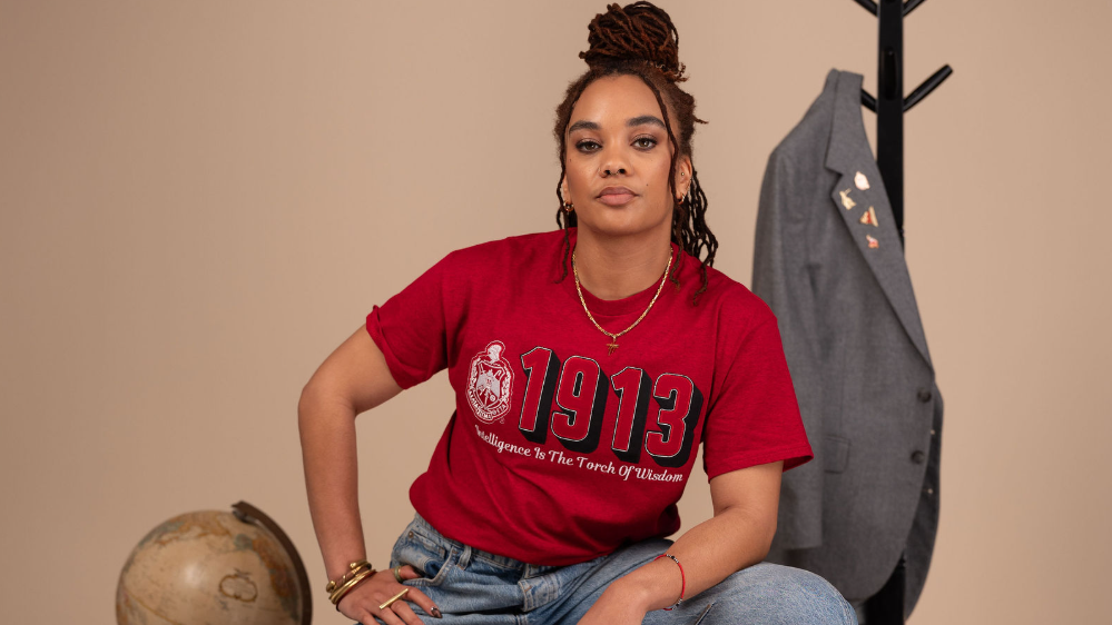 A woman sits on a metal stool wearing a red graphic T-shirt with '1913' and 'Empire State of New York' text, blue jeans, and white sneakers. The setting includes a beige wall, a coat rack with a gray blazer, a globe on a pedestal, and a vintage radio on a patterned rug.